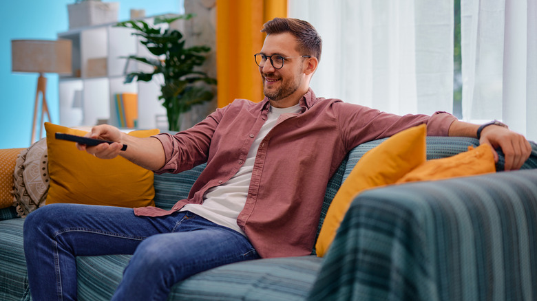 A man in a bright, sun-drenched living room pointing a remote at a television.