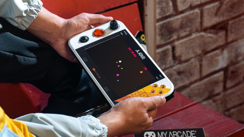 Hands holding a small Atari-themed handheld gaming device with a joystick and button layout.