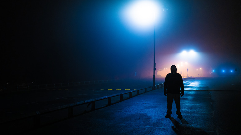 A man in a hoodie standing alone under the bright cone of light from a streetlight at night.