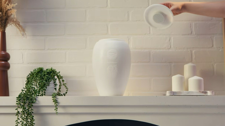 A person lifts the top off the Liquid Death Eternal Playlist Urn which is displayed on a white mantel surrounded by candles and flowers.