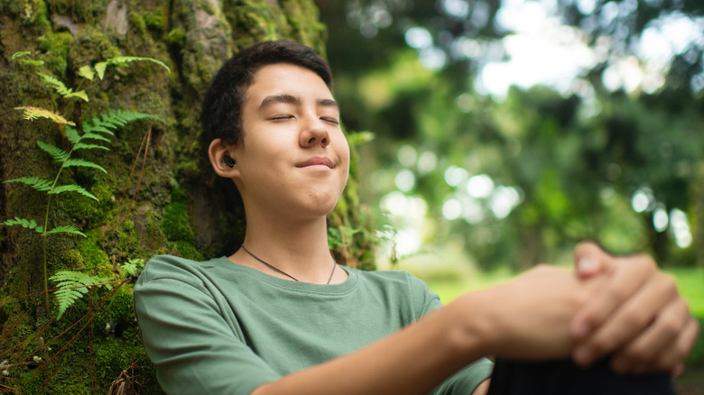 A young person relaxing outdoors against a tree while wearing modern wireless earbuds.