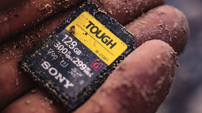 A close-up of a hand covered in sand holding a Sony Tough series SD card to demonstrate its environmental resistance.