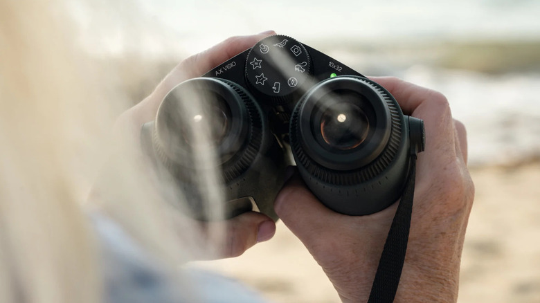 A birdwatcher holding Swarovski AX Visio smart binoculars in an outdoor natural setting.