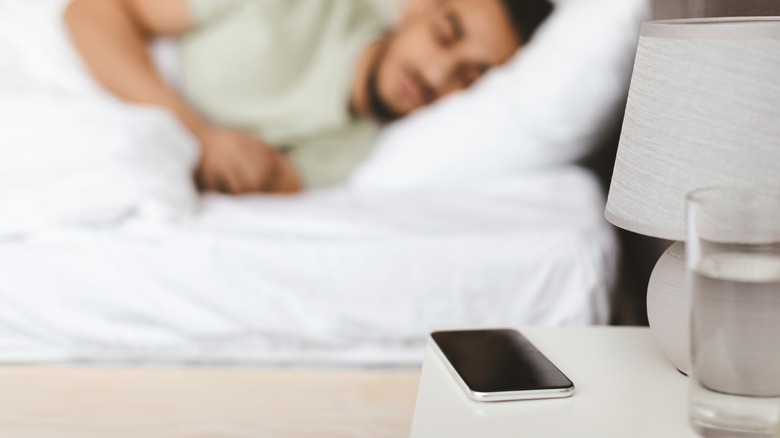 A smartphone resting on a bedside table with a person sleeping in the background.