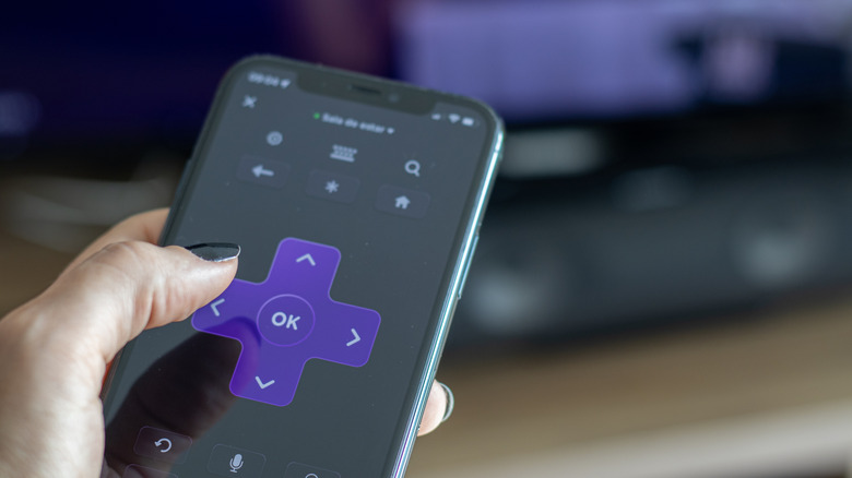 A person using the Roku mobile app on their smartphone to control a television screen in the background.