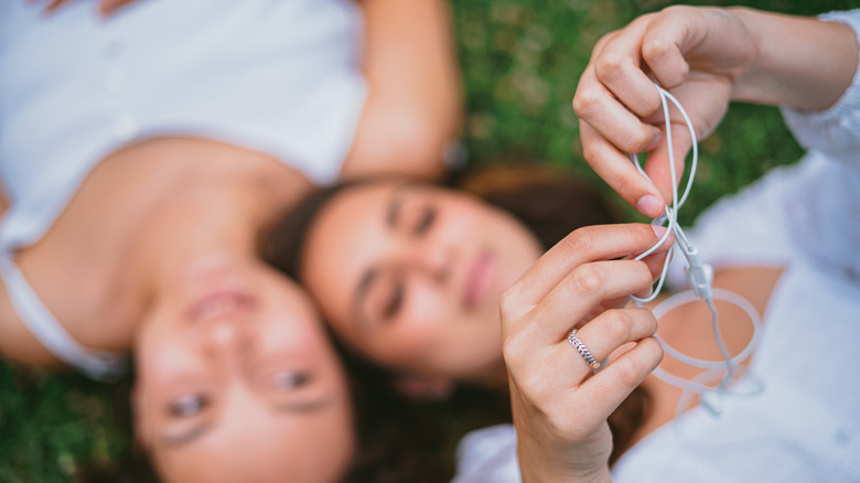 Close-up of white wired earbuds being untangled by two people sitting in the grass.