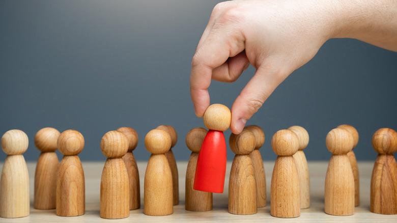A hand selecting a distinct red wooden figure from a line of neutral figures.