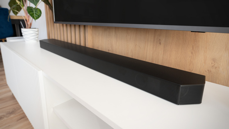 A compact grey soundbar sitting on a white TV stand below a flat-screen television.