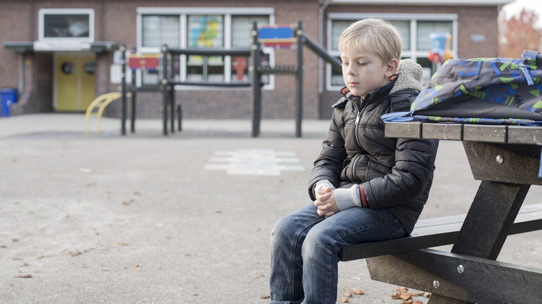 A young boy sitting alone on a bench in a schoolyard environment.