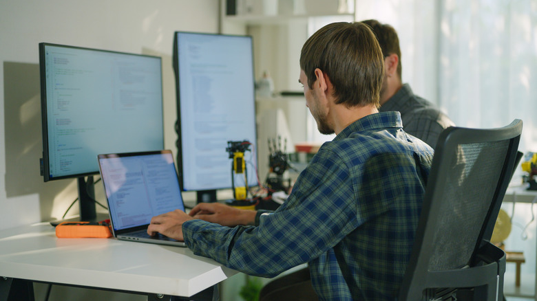 A laptop connected to a larger secondary monitor on a desk in a shared office space.
